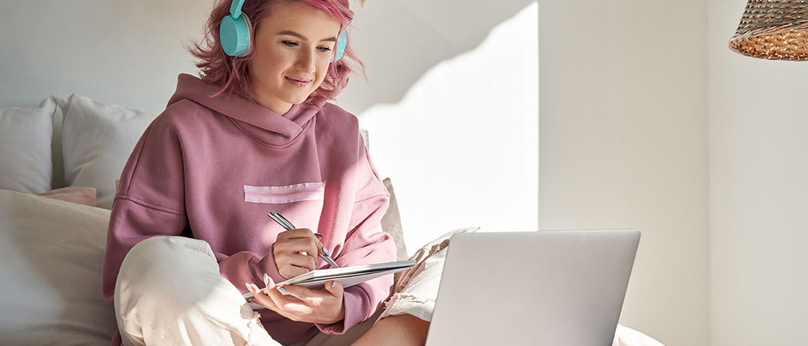 A smiling young person with pink hair, dressed in a hooded sweatshirt and wearing headphones, sits on her bed and takes notes on her laptop.
