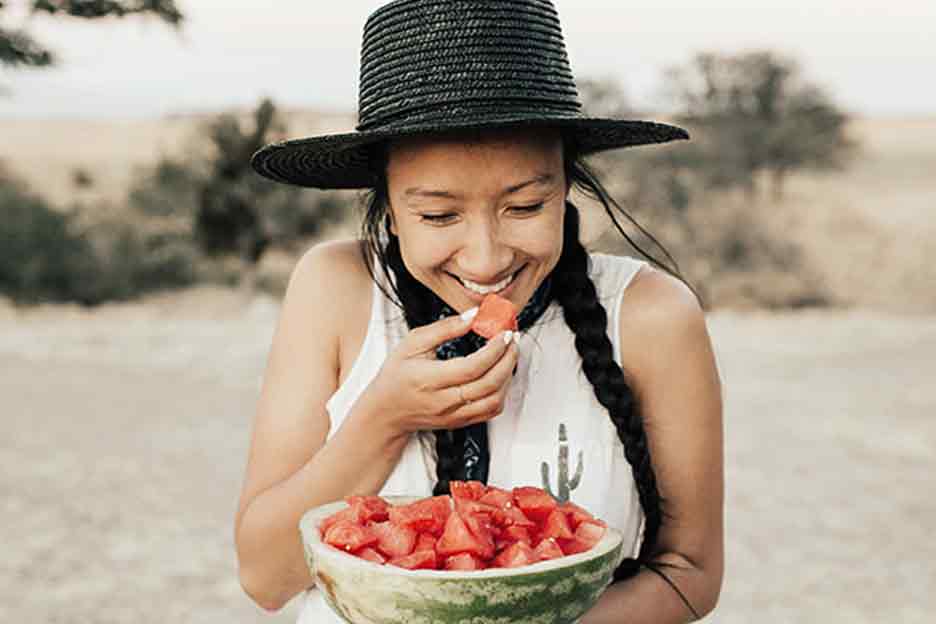 A smiling young woman wearing shorts and a camisole eats watermelon outside on a beautiful summer day.