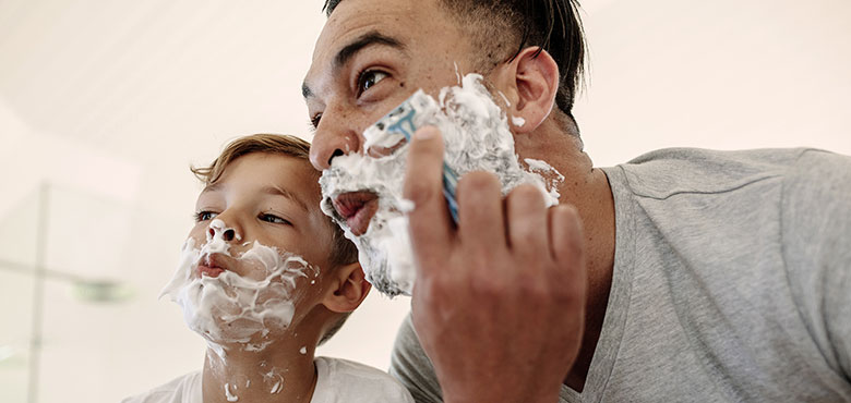 A father and his young son look at their shaving cream–covered faces in the bathroom mirror.
