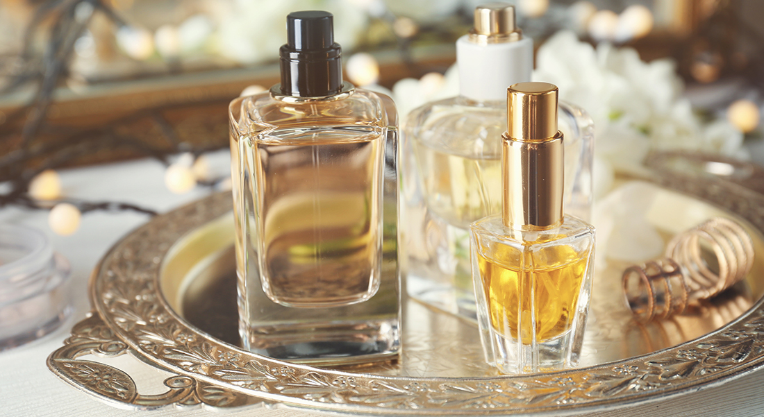 Trois flacons de parfums sont posés sur un comptoir blanc dans un plateau métallique décoratif, avec un miroir et une guirlande de petites lumières blanches en arrière-plan. 