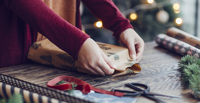 Gros plan sur les mains d'une femme en train d'envelopper un cadeau des Fêtes, entourée de ciseaux et de papier d'emballage, avec un sapin illuminé derrière elle. 