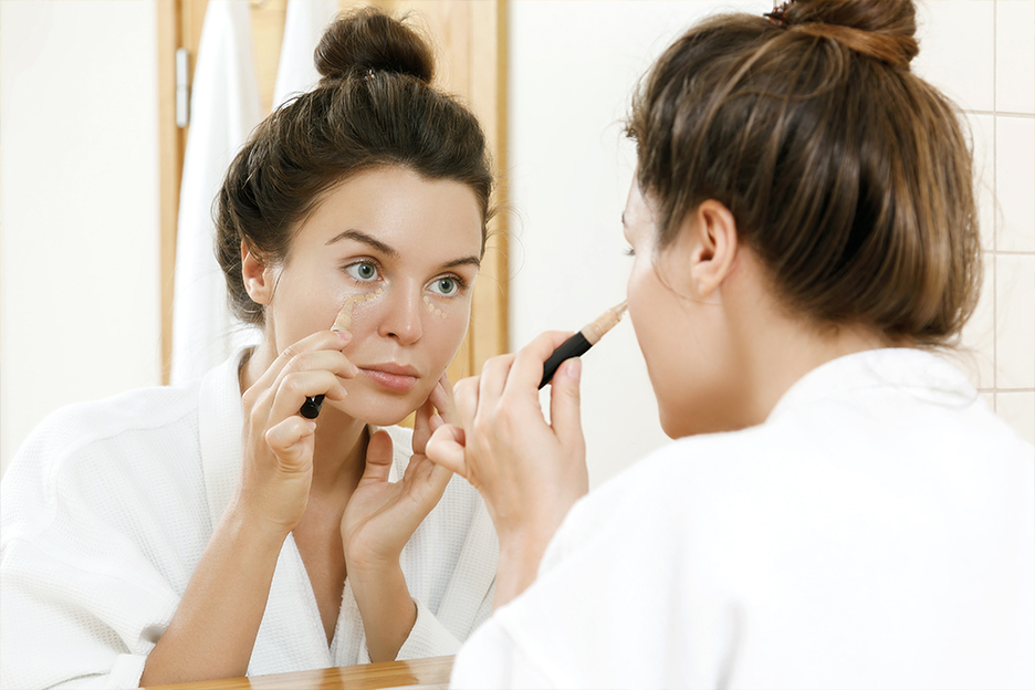 A woman in front of a mirror applies concealer under her eyes.