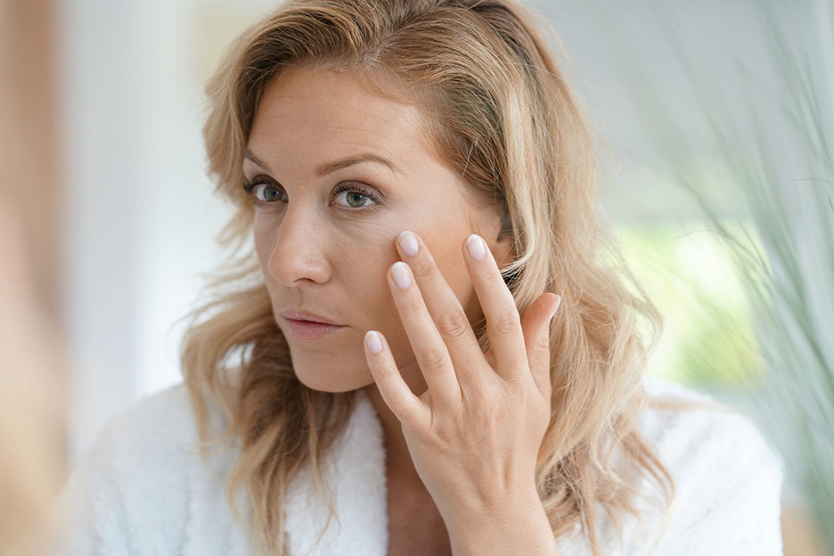 Woman with blonde hair noticing signs of stress on her face as she looks in the mirror.