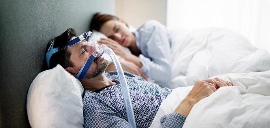 Homme portant un masque CPAP pendant son sommeil pour traiter son apnée du sommeil, avec sa partenaire endormie à ses côtés.